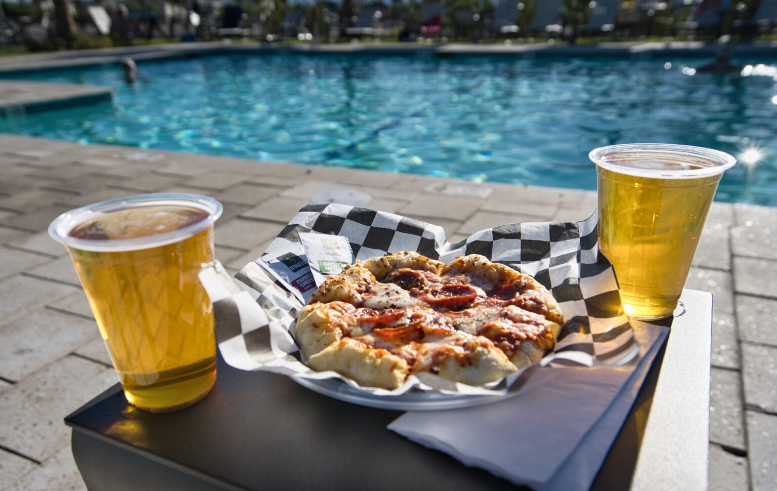 A pepperoni pizza and two cups of beer on a table by a swimming pool, with people swimming in the background.