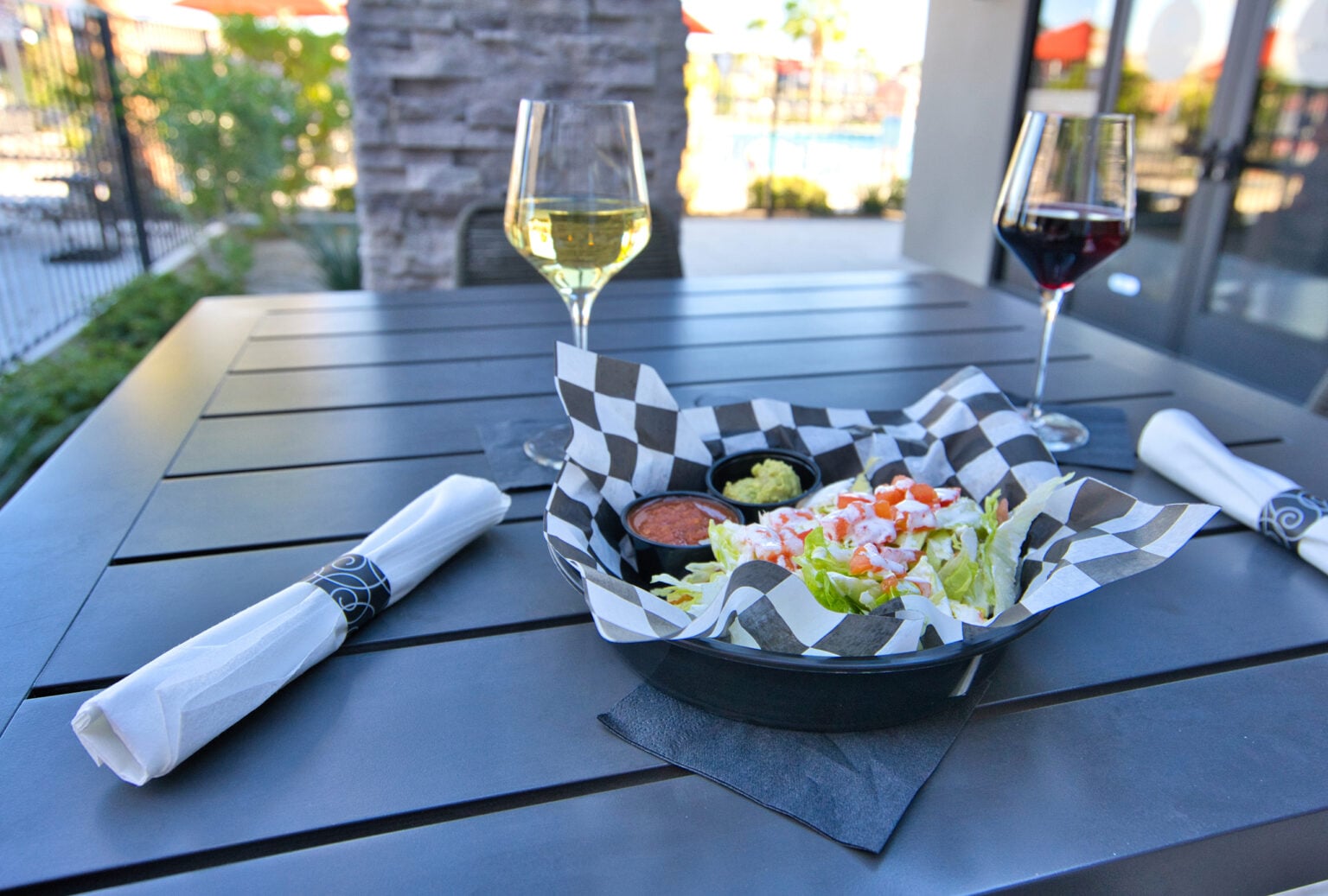 A plate of taco salad with salsa and guacamole sits on an outdoor table alongside two glasses of wine and neatly wrapped napkins.