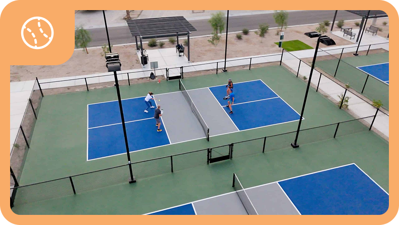 Aerial view of two pickleball courts with four people playing a game; surrounding area includes benches, shade structures, and landscaping.