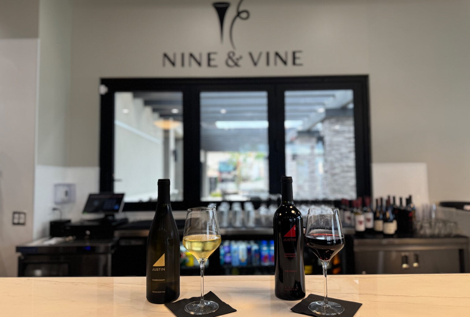 Two wine bottles and filled glasses—one white, one red—are displayed on a bar counter at Nine & Vine, with shelves of wine in the background.
