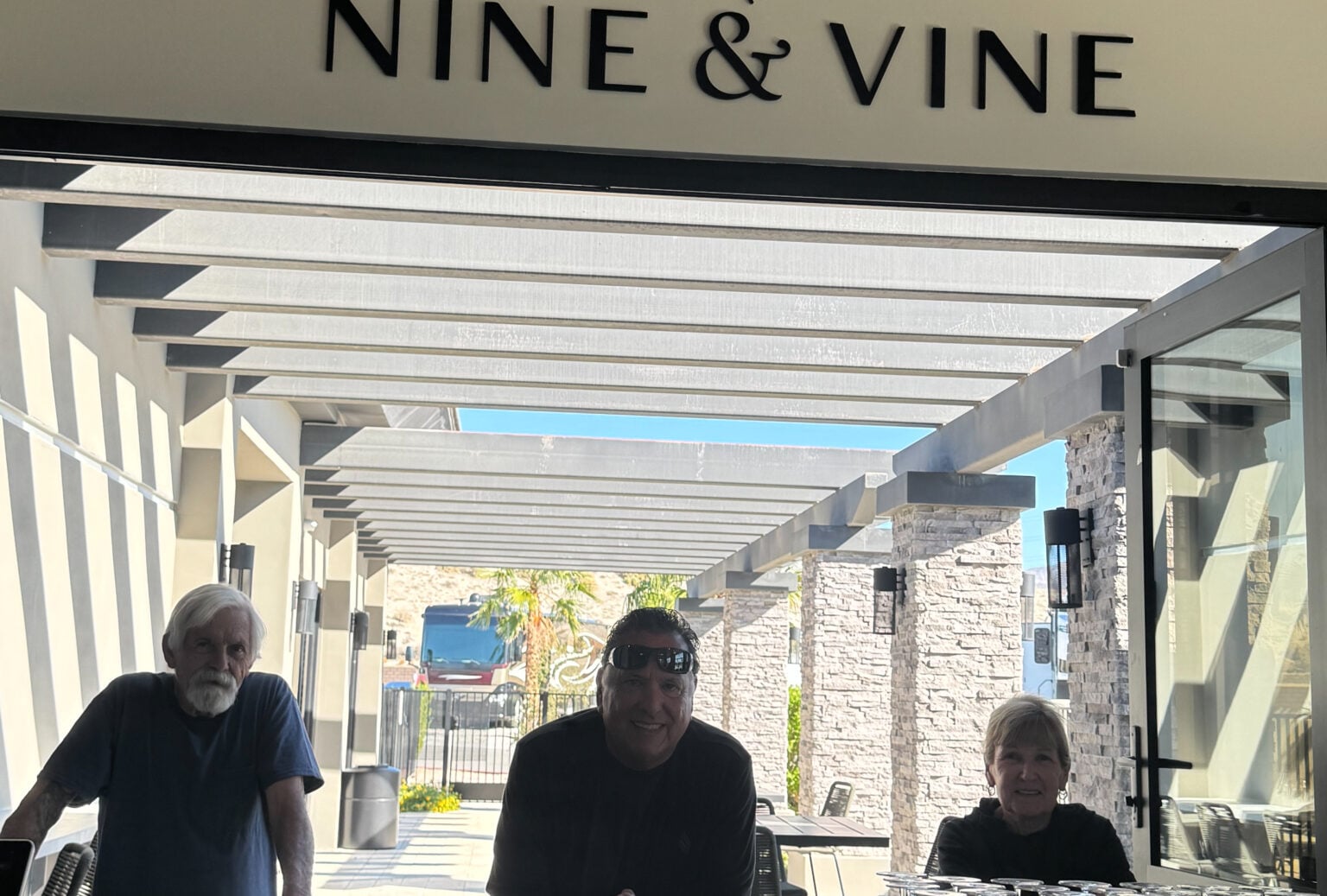Three people stand and sit beneath a pergola at the entrance of a place called "NINE & VINE," with sunlight streaming in from behind them.