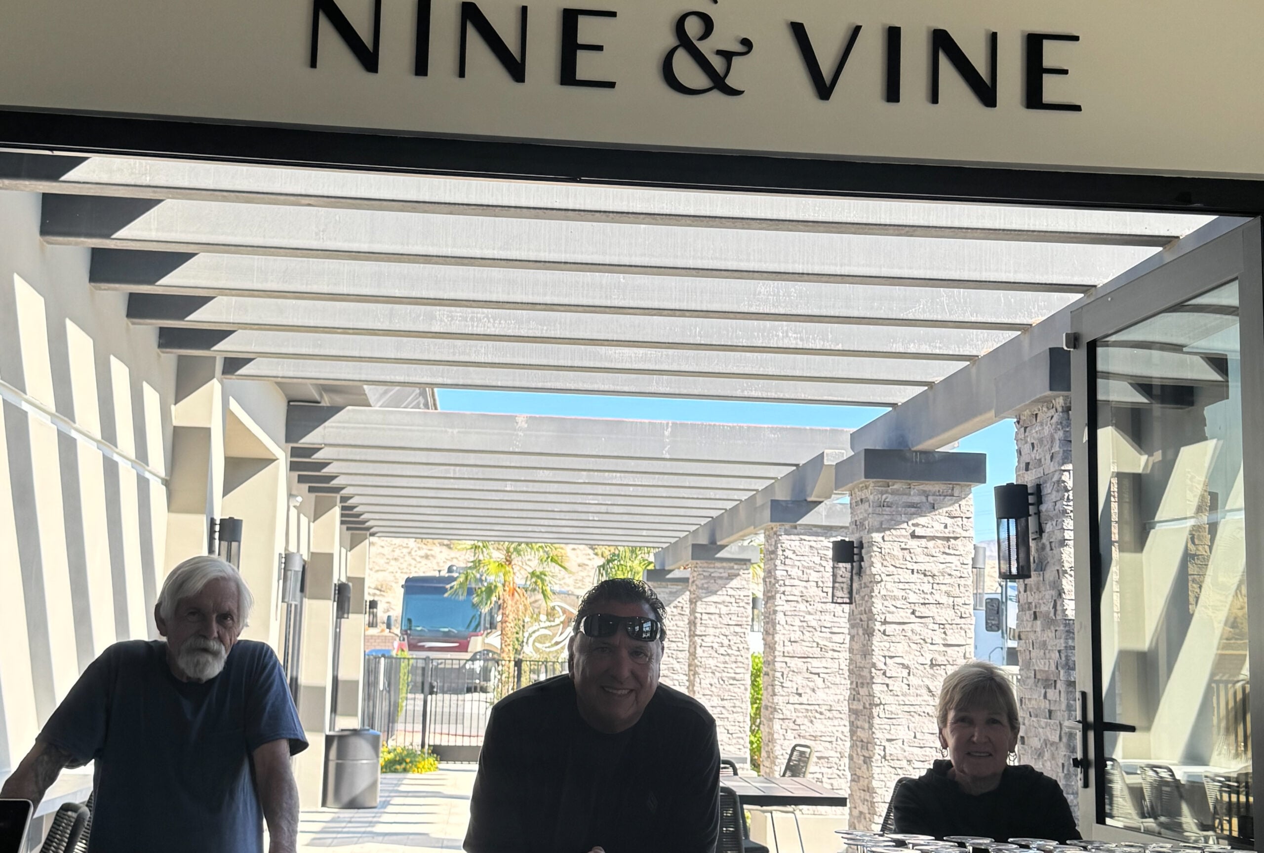 Three people stand and sit beneath a pergola at the entrance of a place called "NINE & VINE," with sunlight streaming in from behind them.