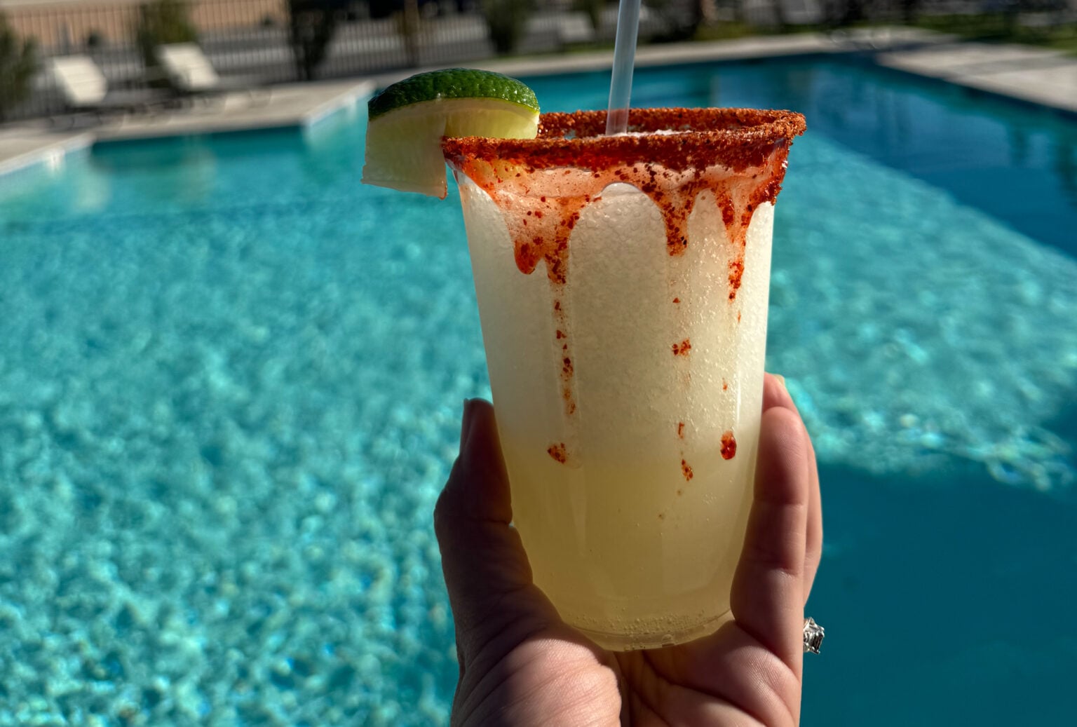 A hand holds a frozen cocktail with a chili powder rim, lime wedge, and straw in front of a swimming pool on a sunny day.