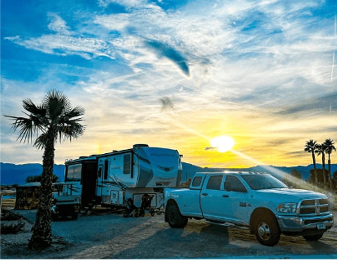 A white pickup truck is parked in front of a travel trailer at a campsite, with palm trees and a bright sunset in the background.