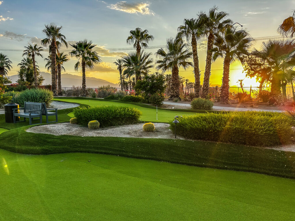 Palm trees and shrubs surround a well-manicured golf putting green at sunset, with benches and mountains visible in the background.