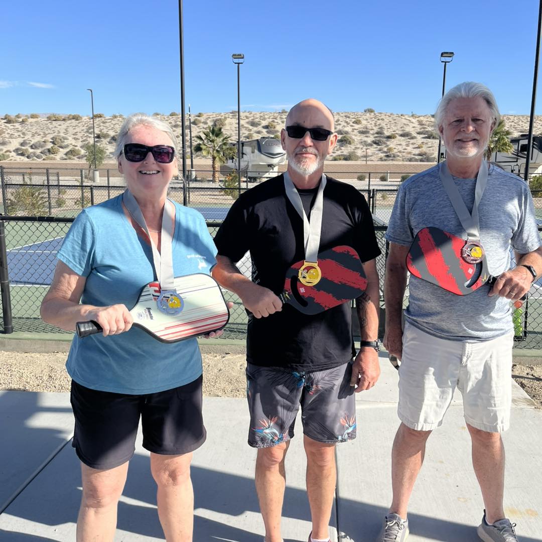 Three older adults stand outdoors on a sunny day, each holding a pickleball paddle and wearing a medal around their neck. A desert landscape and fenced courts are visible in the background.