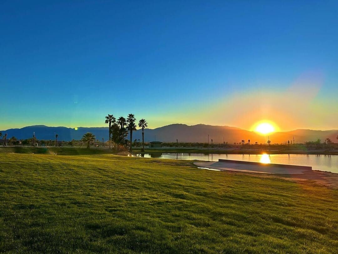 Sunset over a grassy field with palm trees, a pond, and mountains in the background under a clear blue sky.