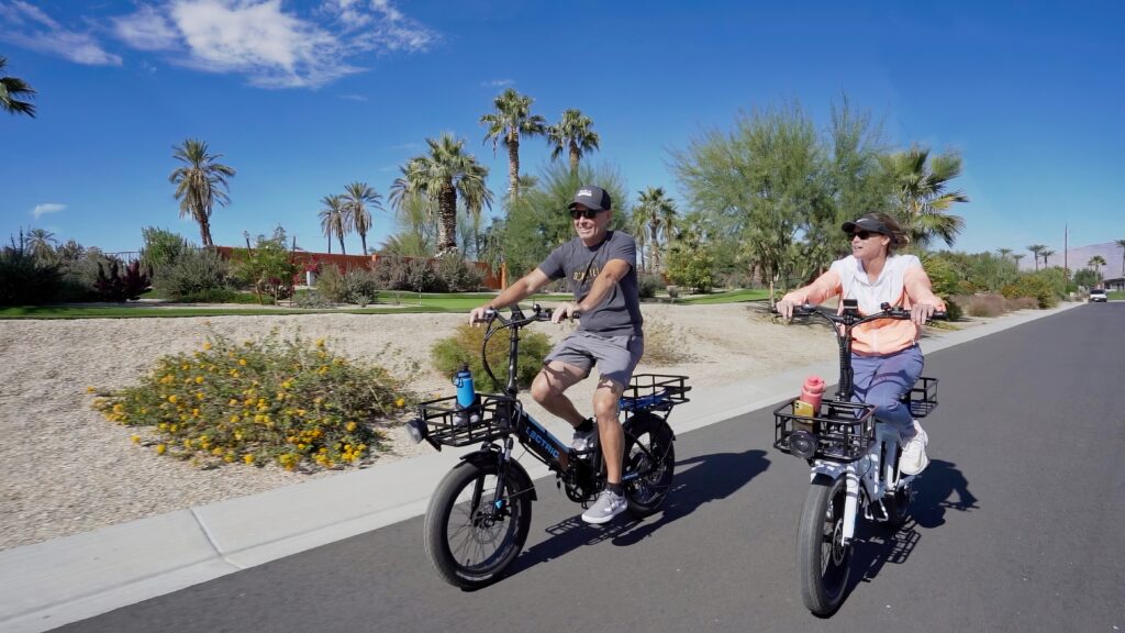 Two people ride electric bicycles on a sunny suburban street lined with palm trees and desert landscaping.