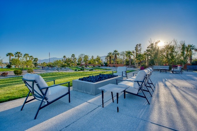 Outdoor patio with cushioned chairs and a fire pit overlooks a grass lawn with palm trees, under a clear blue sky and bright sun.