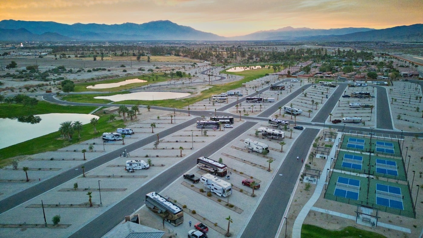 Aerial view of an RV park with lined parking spaces, several RVs, tennis courts, ponds, palm trees, and mountains in the background at sunset.