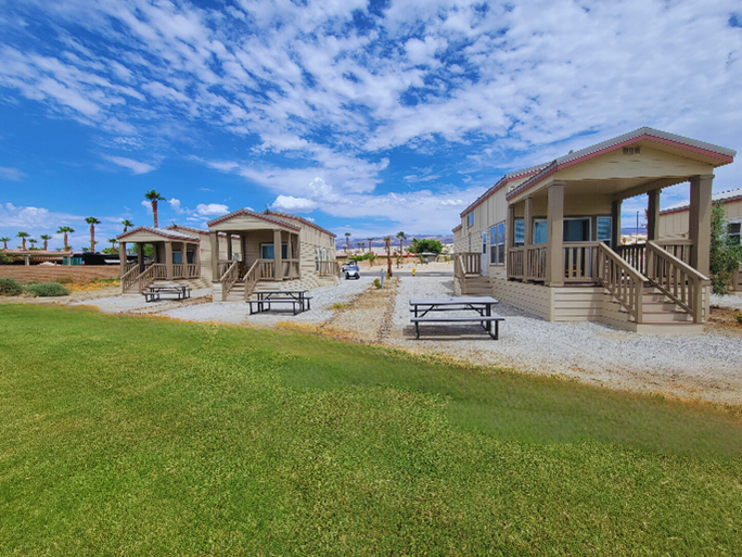 Three small cabins with front porches and picnic tables stand in a row on gravel plots, surrounded by grass and under a partly cloudy sky.