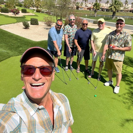 A group of six men pose and smile on a sunny mini golf course, with putters and a green visible in the background.
