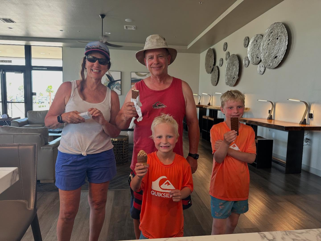 Two adults and two children stand indoors, each holding an ice cream treat. The adults wear hats and sunglasses, and all are casually dressed. The background shows modern decor and wall art.