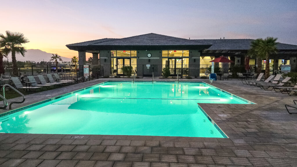 Outdoor swimming pool with clear water, surrounded by lounge chairs and palm trees, in front of a modern building at sunset.