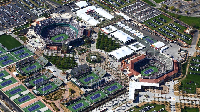 Aerial view of a large tennis complex with multiple courts, two main stadiums, surrounding parking lots, and spectator areas.