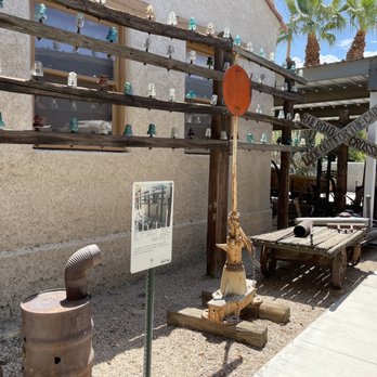 Outdoor museum display featuring vintage telegraph poles with glass insulators, a historical sign, old equipment, and a wooden cart on a gravel area beside a building.