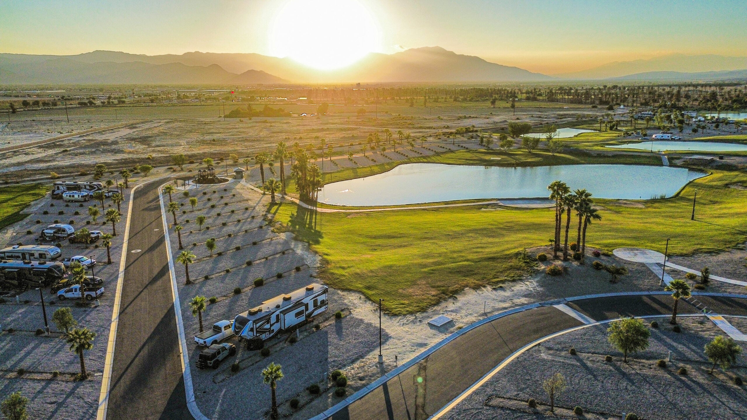 Aerial view of an RV park with several RVs parked near a pond, palm trees, and mountains in the background at sunset.