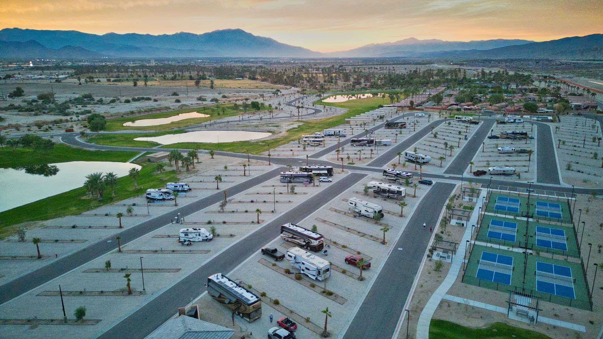 Aerial view of an RV park with rows of parked RVs, several pickleball courts, small ponds, palm trees, and mountains in the background at sunset.