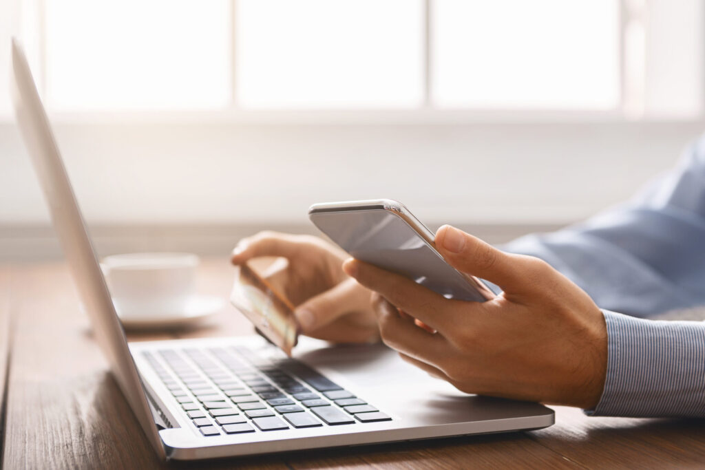 Person using a laptop while holding a credit card in one hand and a smartphone in the other, likely performing an online transaction at a desk.