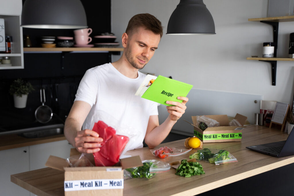 A man stands in a kitchen reading a recipe card, with meal kit ingredients and boxes arranged on the counter in front of him.