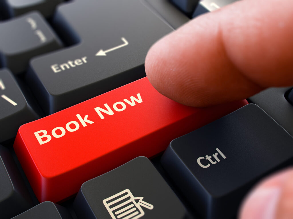 A finger presses a red keyboard key labeled "Book Now" located between the Enter and Ctrl keys.