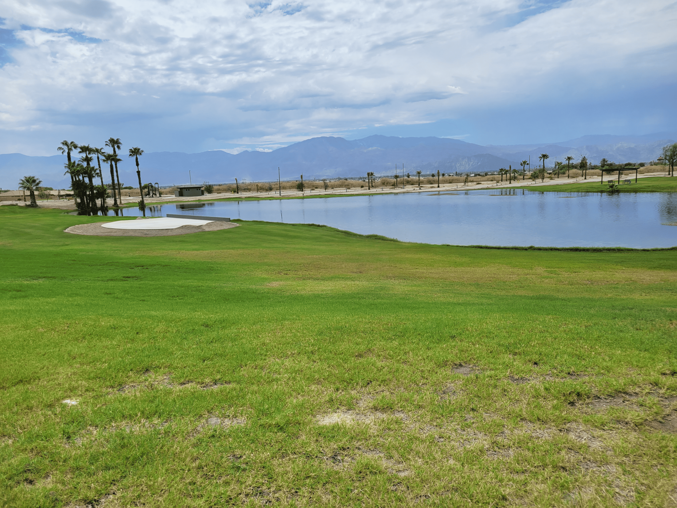 A golf course with a sand bunker, grassy fairway, and a pond, with mountains and cloudy sky in the background.