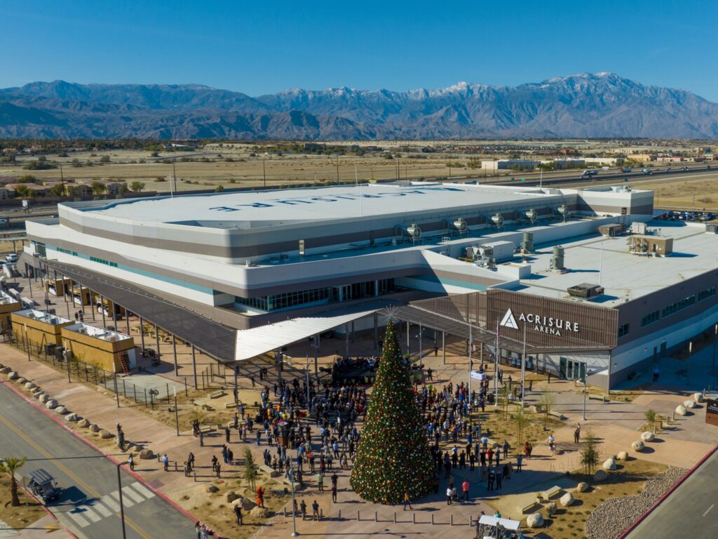 Aerial view of Acrisure Arena with a large decorated Christmas tree and a crowd gathered outside, mountains visible in the background.