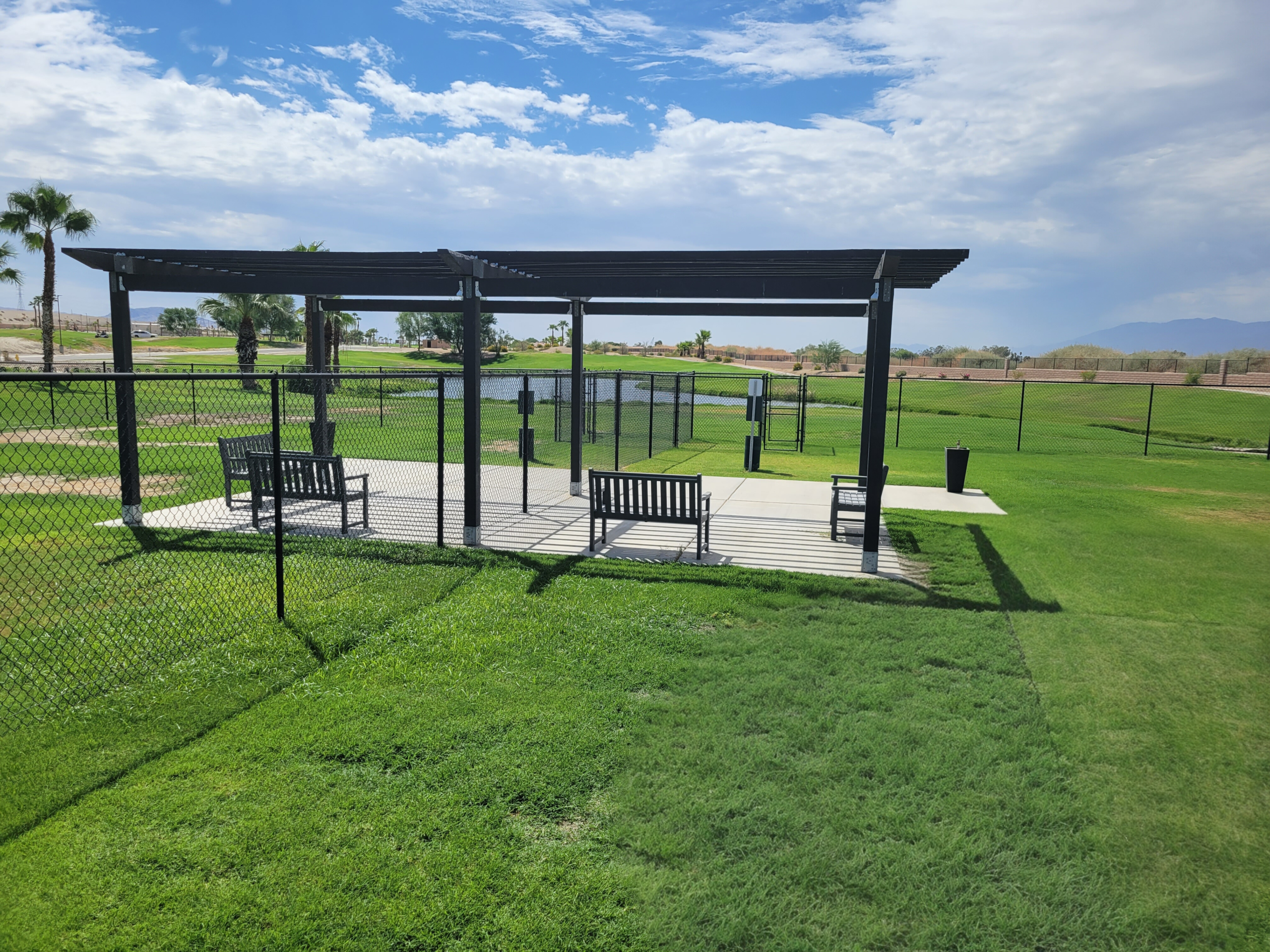A fenced grassy area with benches and a shaded pergola, likely part of a park or recreational space, under a partly cloudy sky.