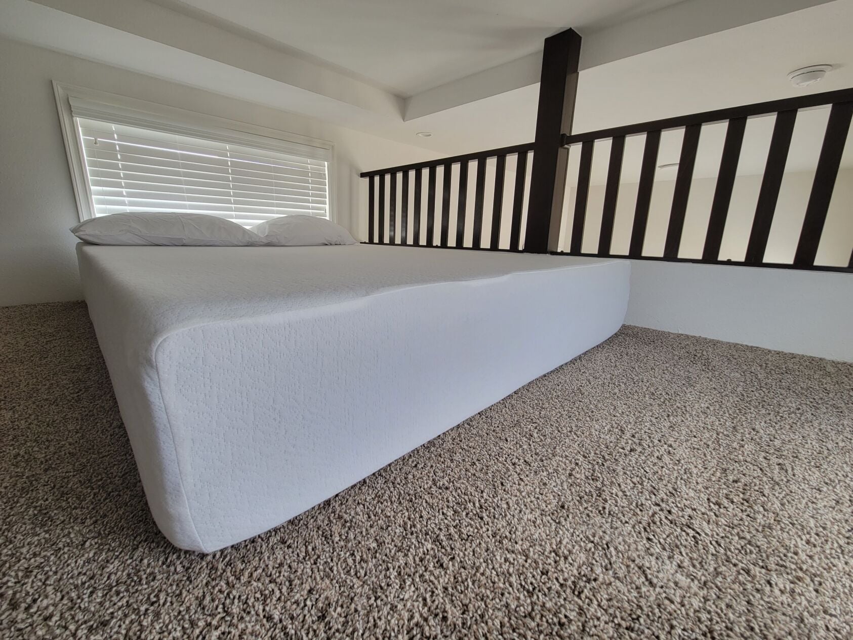A mattress with white bedding and two pillows sits on a carpeted floor next to a railing and a window with closed blinds.