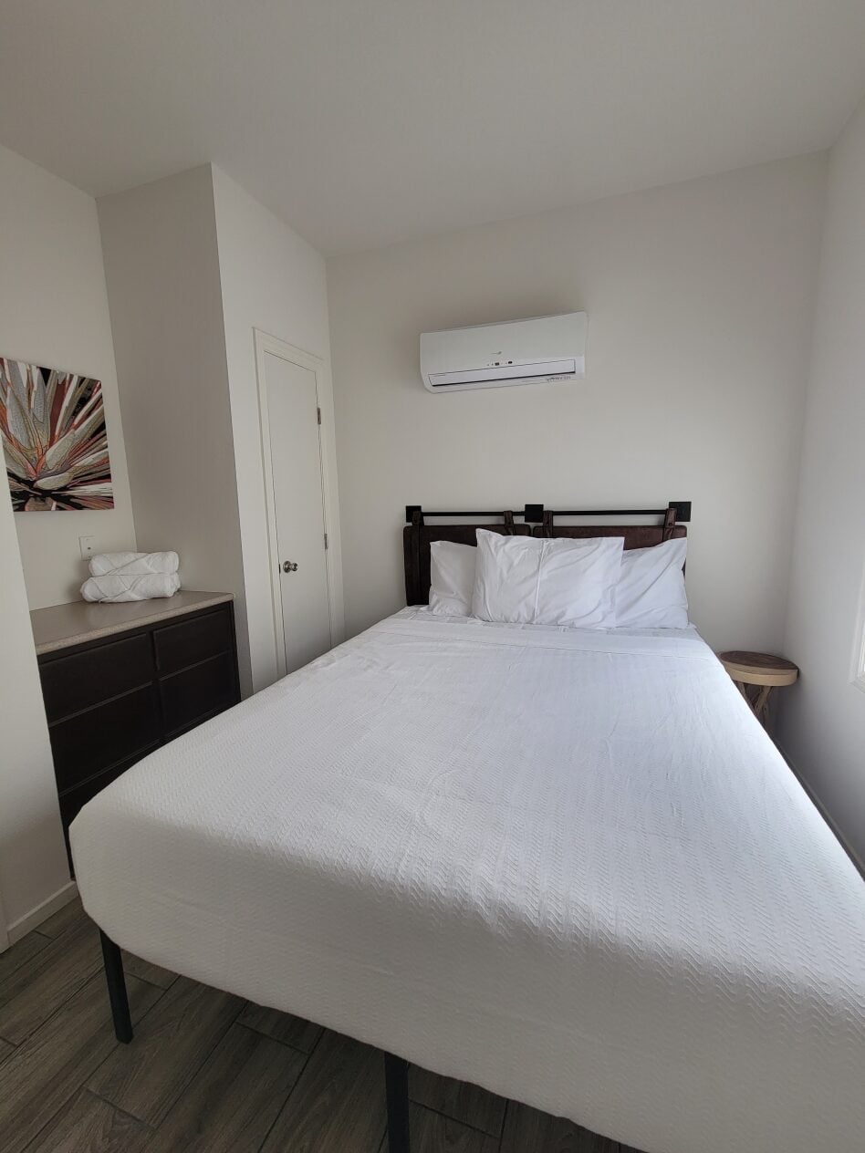 A neatly made bed with white linens is centered in a small, white-walled bedroom with an air conditioner above the headboard and a dresser with folded towels on the left.