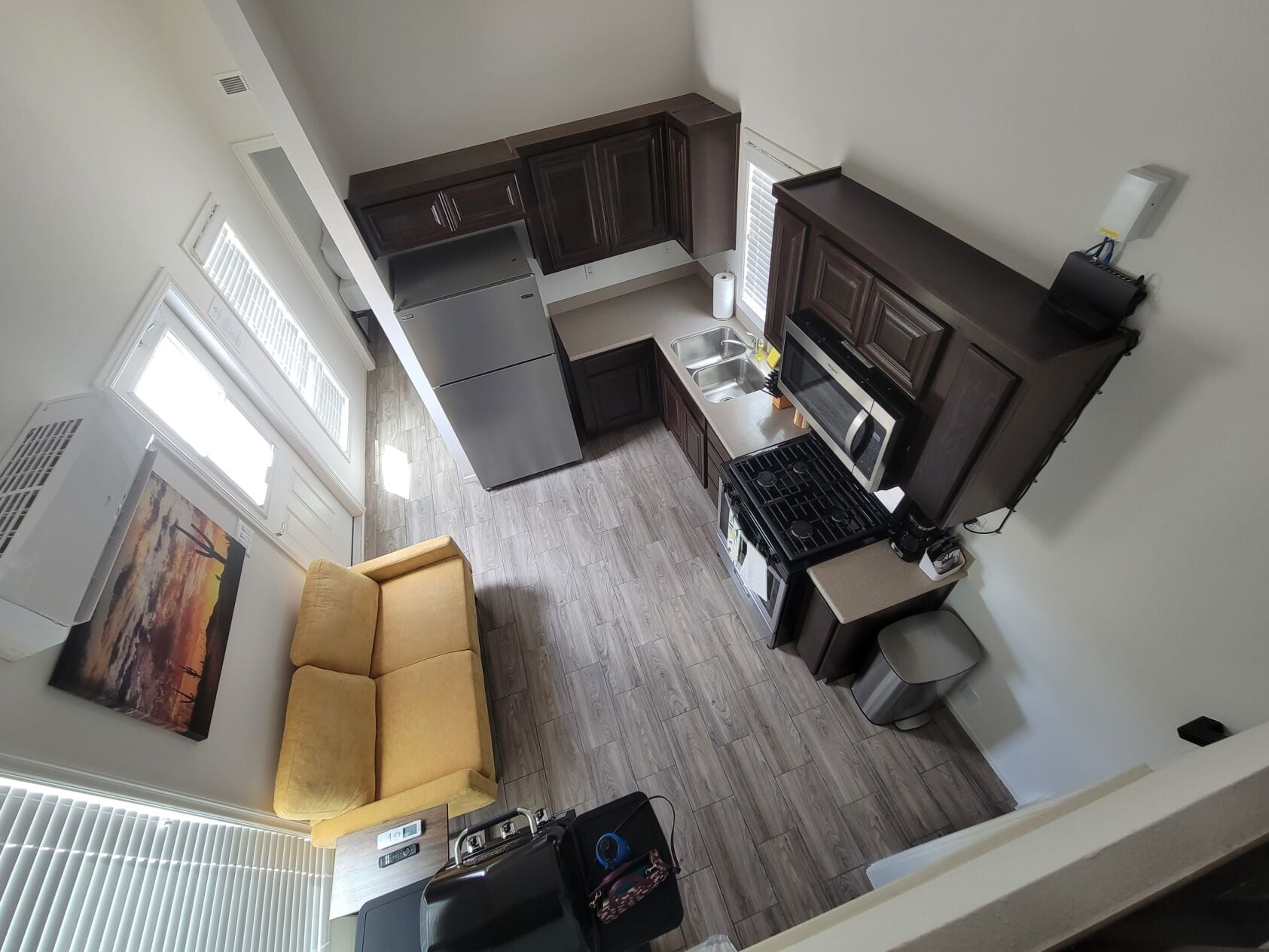 Overhead view of a compact kitchen and living area with dark cabinets, stainless steel appliances, a yellow sofa, and a landscape painting on the wall.