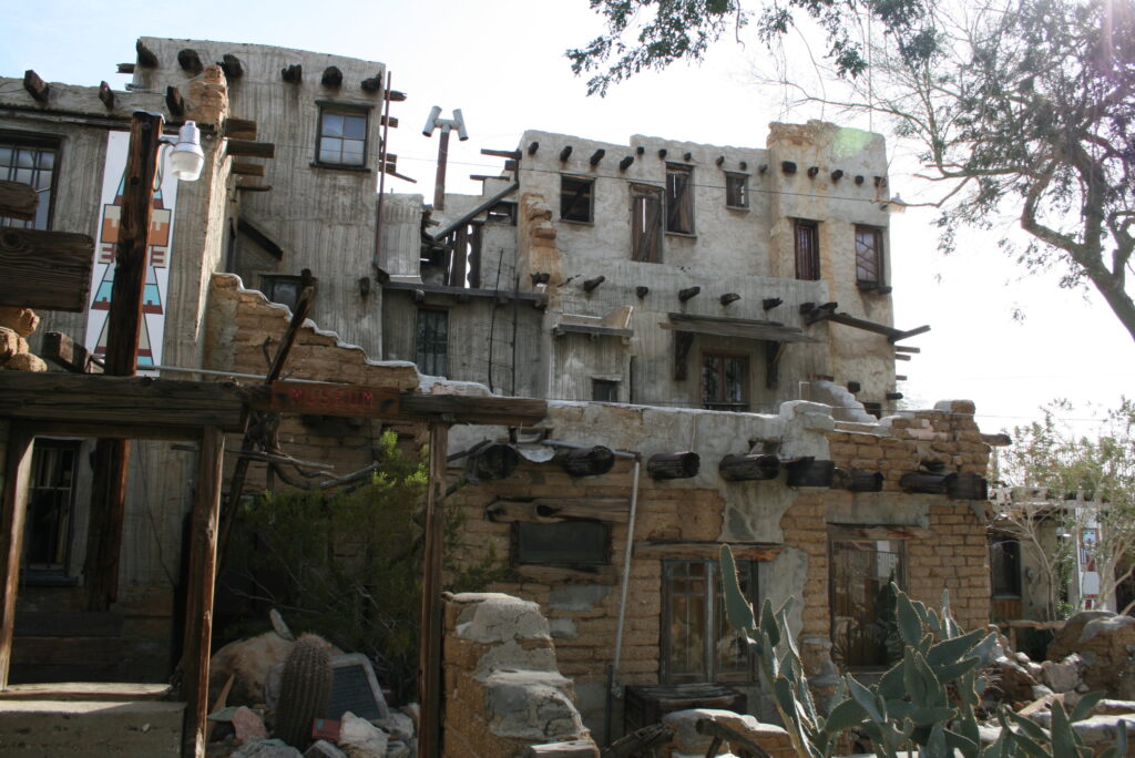 A multi-story adobe-style building with weathered walls, wooden beams, small windows, and desert plants in front, under a clear sky.