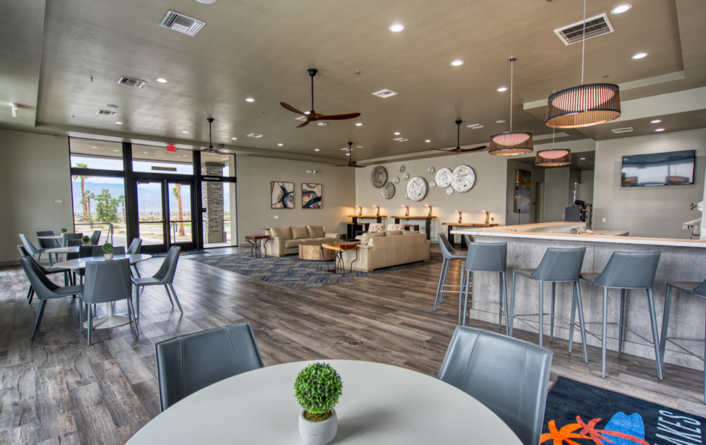 Modern lounge area with gray chairs, round tables, a bar counter, sofas, wall clocks, and ceiling fans, featuring large windows and wood flooring.