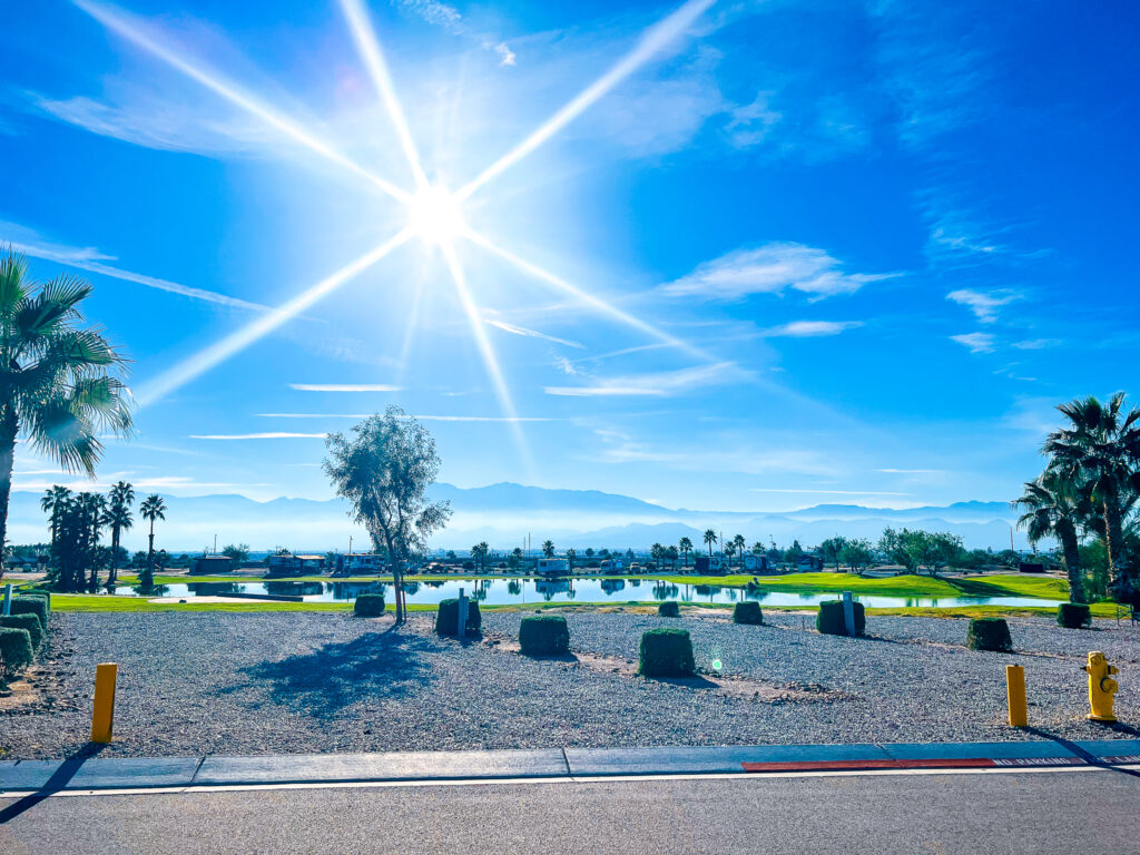 A bright sun shines over a landscaped golf course with palm trees, a pond, and distant mountains under a clear blue sky.