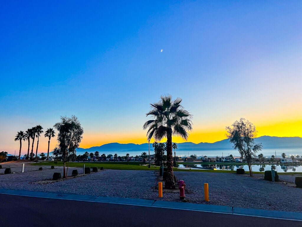 View of palm trees, a parking area, and a body of water with mountains in the background at sunset, with a crescent moon visible in the clear sky.