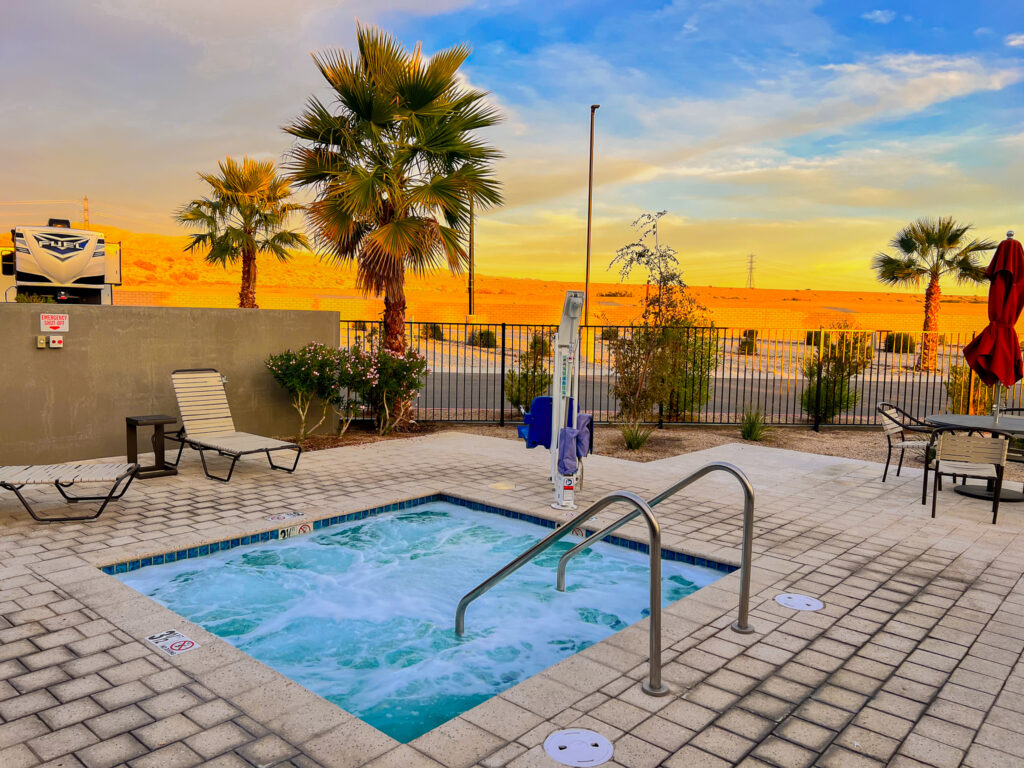 Small outdoor hot tub with bubbling water, surrounded by lounge chairs and palm trees, set against a sunset sky and desert landscape.