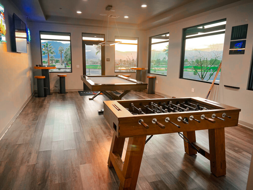 A modern game room with a foosball table, pool table, tall tables with stools, large windows, and wood flooring, with mountains visible outside.