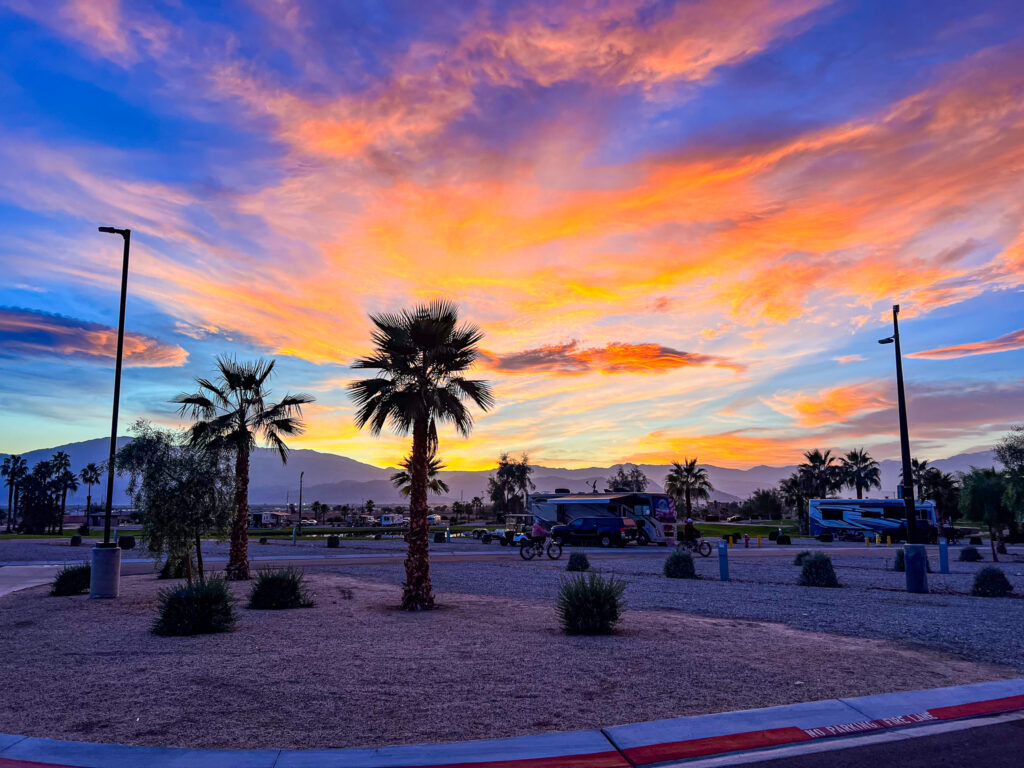 A vibrant sunset sky with pink and orange clouds over a desert RV park, palm trees, and mountains in the background.