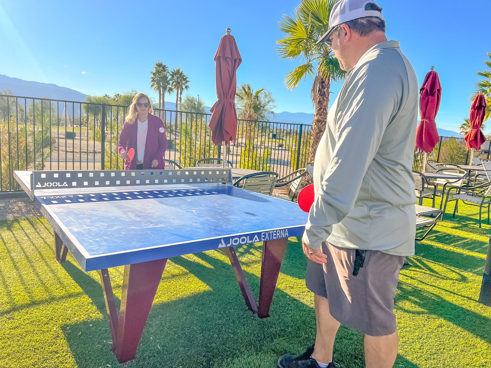 Two people play table tennis outdoors on a sunny day, with palm trees, umbrellas, and a fence in the background.