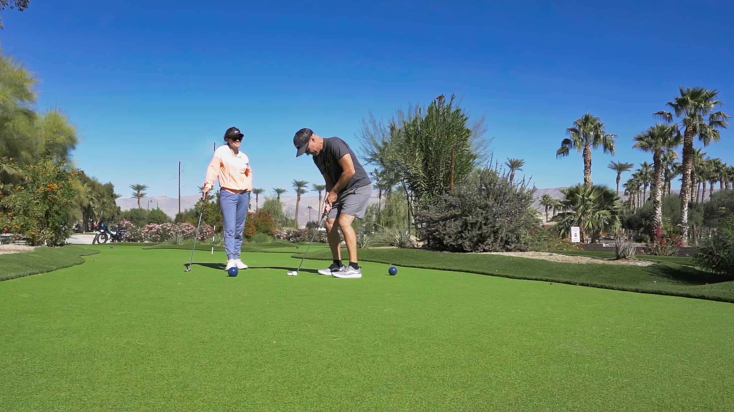 Two people are on a golf course; one is putting while the other watches. The scene features clear skies, palm trees, and well-maintained green grass.