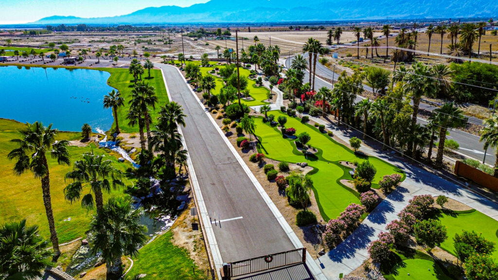 Aerial view of a landscaped park with palm trees, putting greens, a pond, and a road running through the area, with mountains visible in the background.
