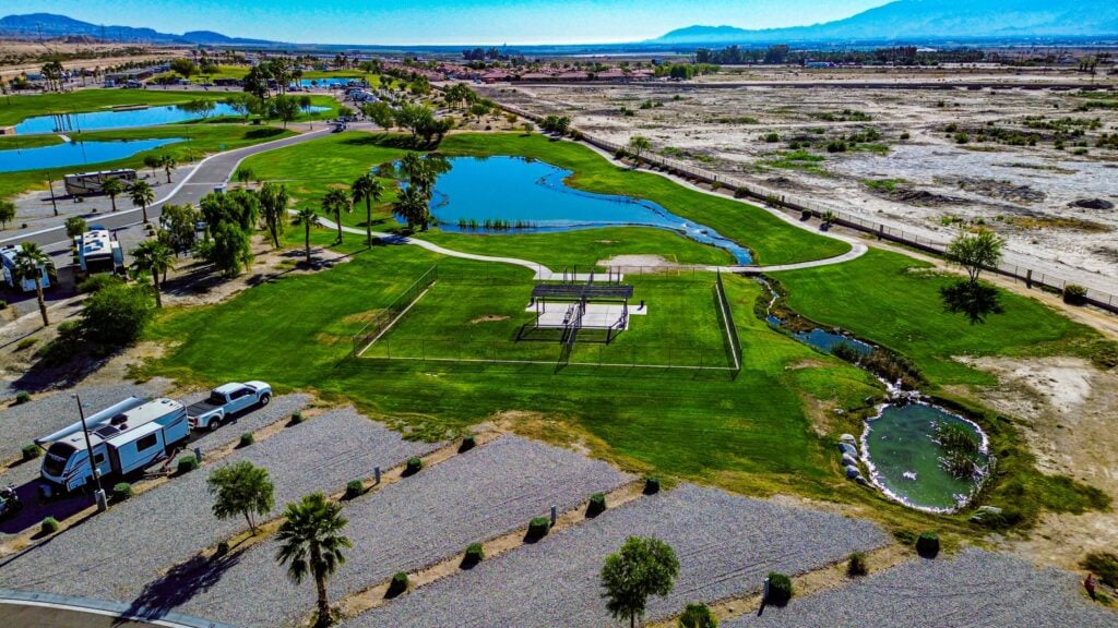 Aerial view of a park with green lawns, a small pond, a gazebo, RVs parked nearby, and a dry landscape in the background under a clear sky.