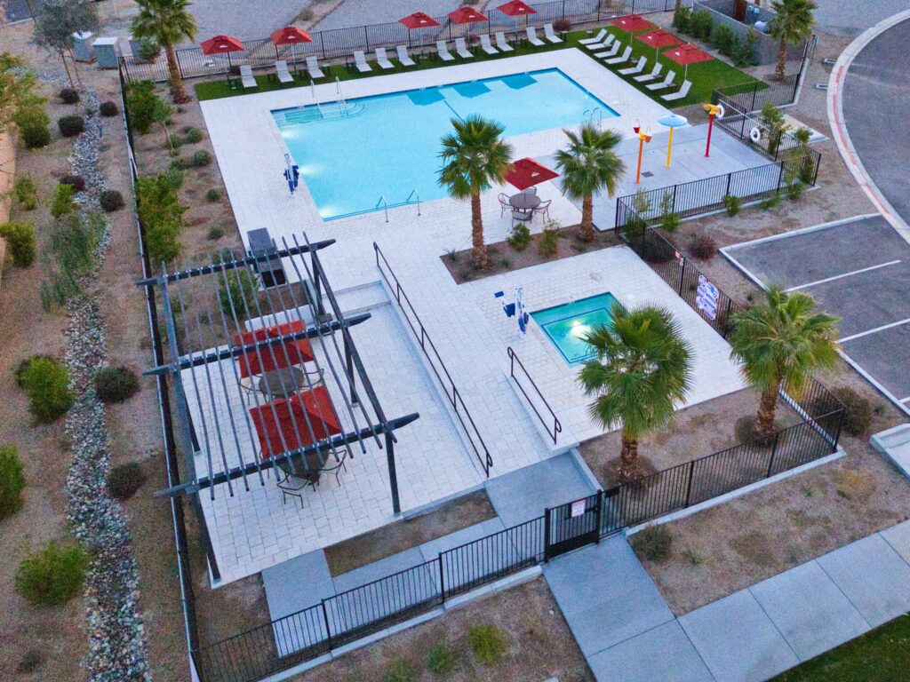 Aerial view of a fenced outdoor pool area with a large swimming pool, a smaller hot tub, lounge chairs, tables with umbrellas, and landscaping with palm trees.