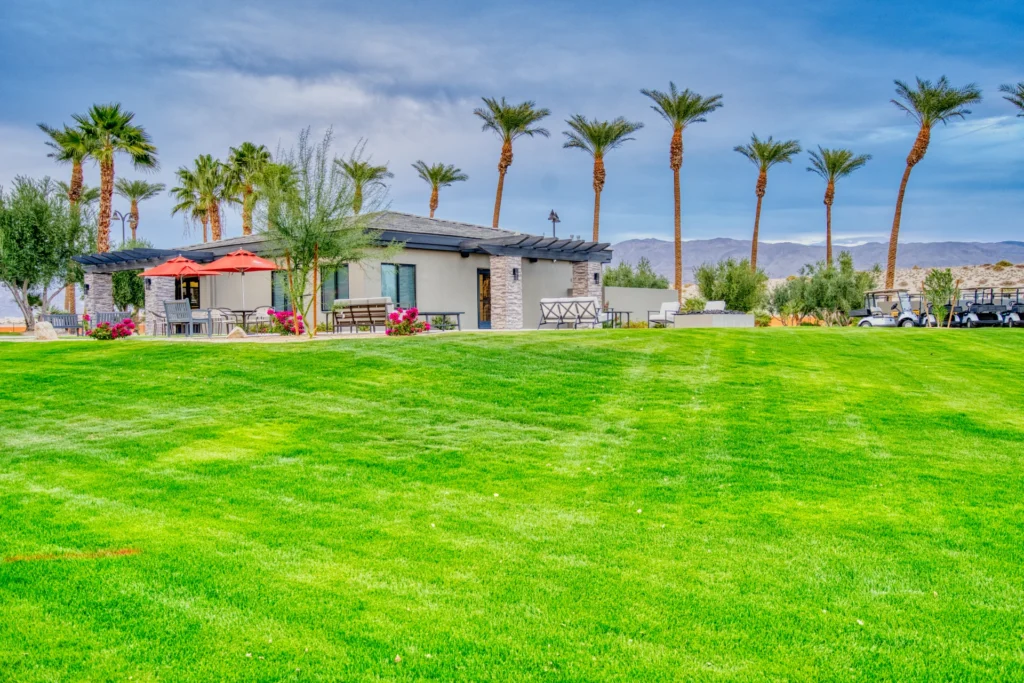 Single-story modern building with red umbrellas on a patio, surrounded by palm trees and a large green lawn under a blue sky with mountains in the background.