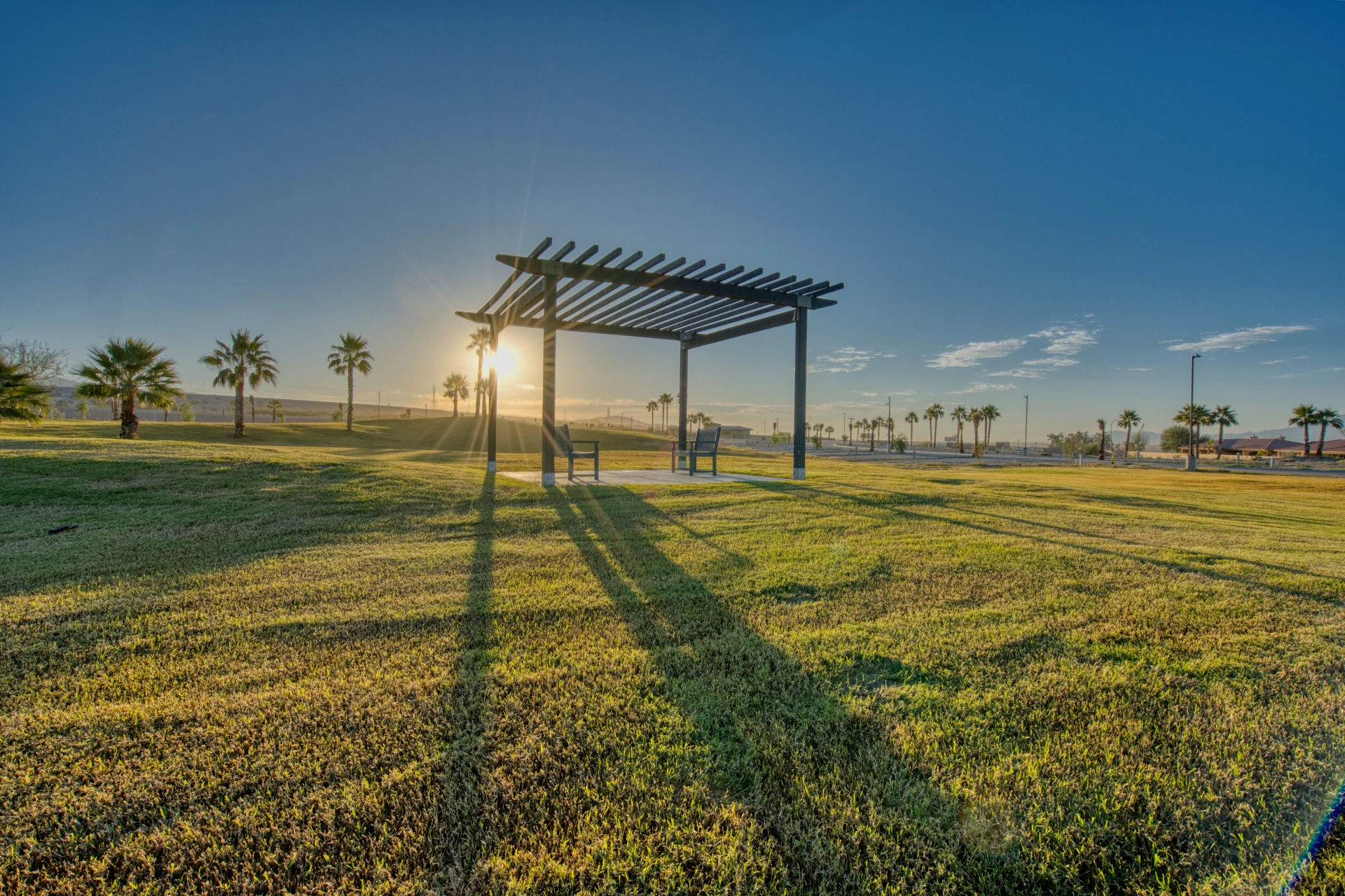 A pergola with two benches stands on a grassy lawn at sunrise, casting long shadows. Palm trees and a clear blue sky are visible in the background.