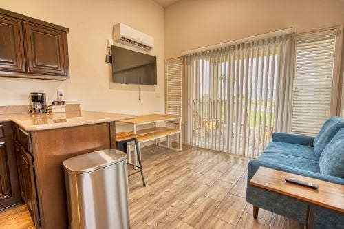 Small living area with a blue sofa, wall-mounted TV, kitchenette, wooden dining table, and sliding glass doors leading to an outdoor patio.