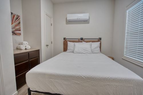 A small bedroom with a neatly made bed, three white pillows, a dresser with rolled towels, a wall-mounted air conditioner, and a window with closed blinds.