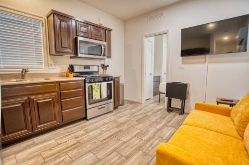 Small kitchen with dark wood cabinets, stainless steel appliances, a mounted TV, and a yellow sofa in a compact living space. A doorway leads to a bathroom.