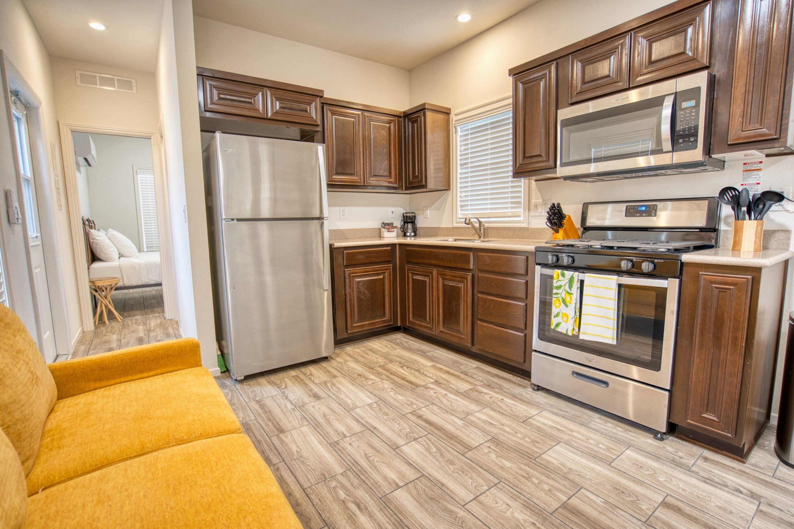 Modern kitchen with stainless steel appliances, dark wood cabinets, beige countertops, and a yellow couch in the foreground. A bedroom is visible through a doorway.