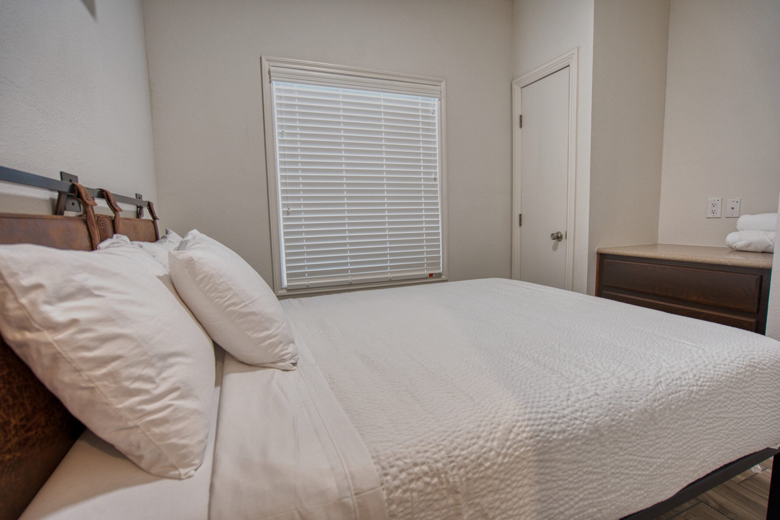 A small bedroom with a neatly made bed, white bedding, a wooden nightstand with towels, and a window with closed blinds.